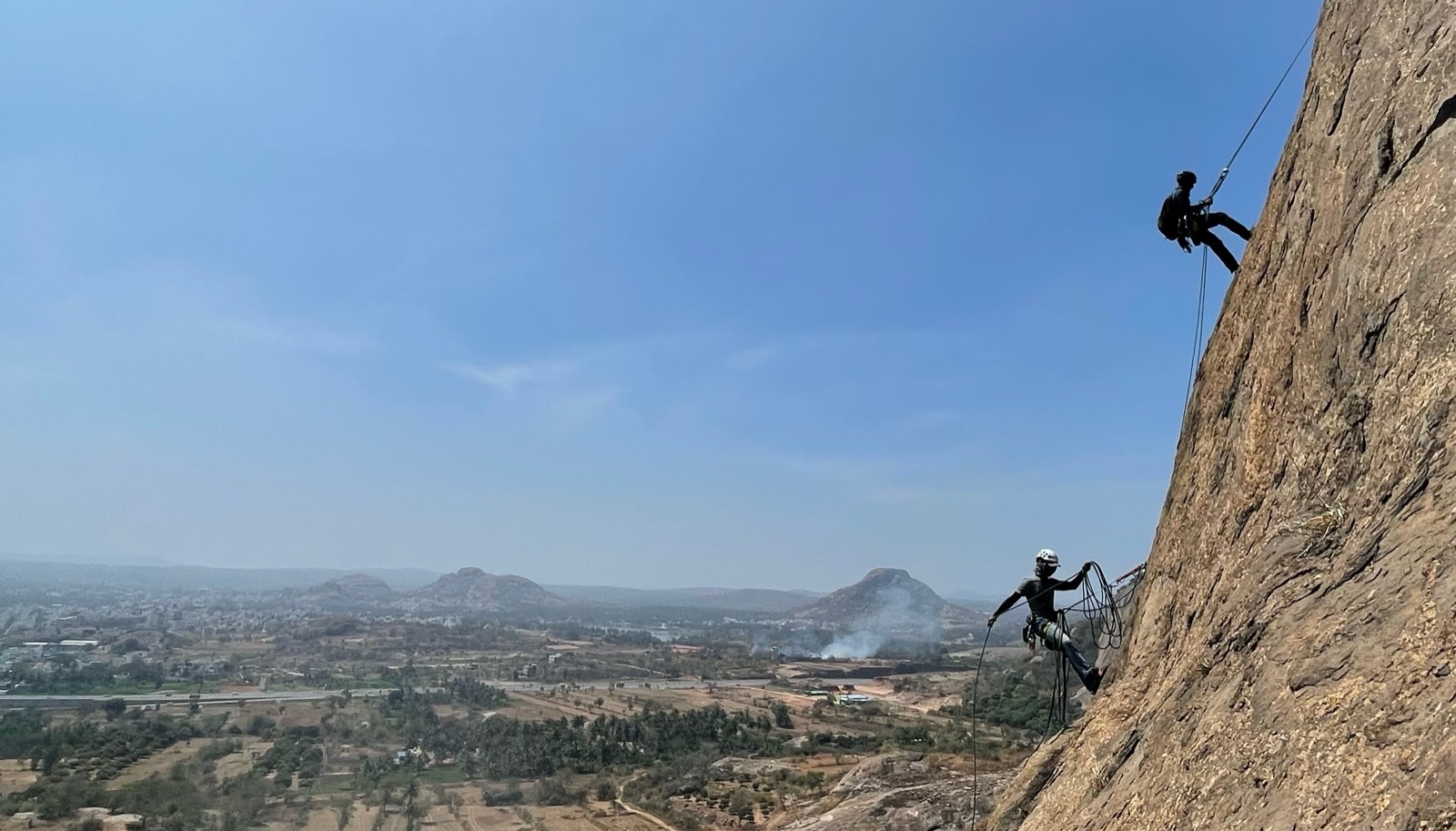Two climbers on a granite dome above the Ramanagaram valley, one
              rappelling and one belaying, with the plain and distant hills
              behind them